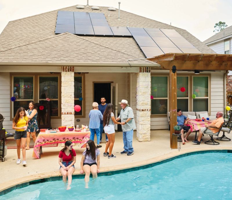 A multigenerational hispanic family enjoying a backyard party at their home with solar panels and a swimming pool.