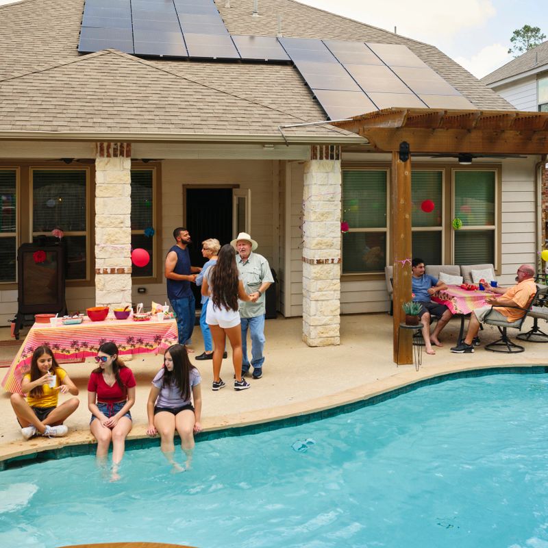 A multigenerational hispanic family enjoying a backyard party at their home with solar panels and a swimming pool.