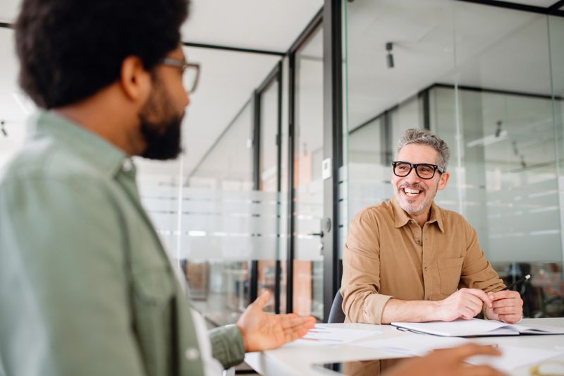 Two male professional colleagues are engaged in a friendly discussion at the office, the older man in glasses displays a warm smile, indicating a positive work environment