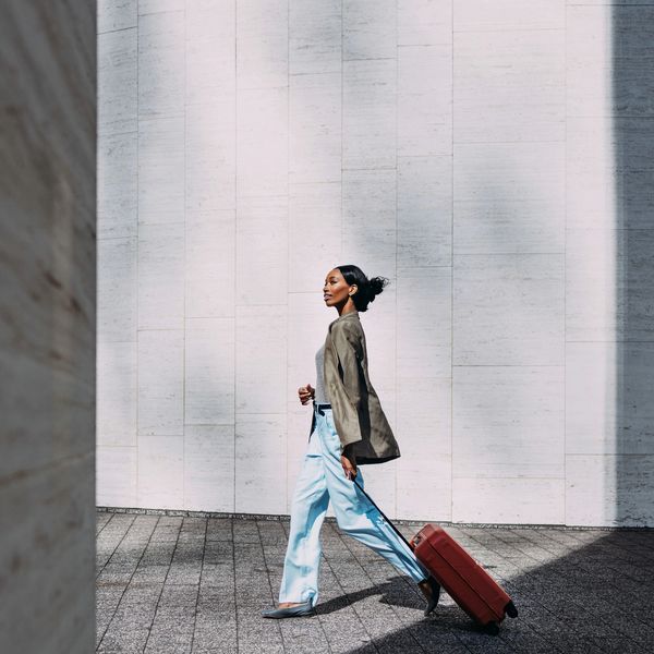 Woman walking with a red suitcase against a modern wall in sunlight.