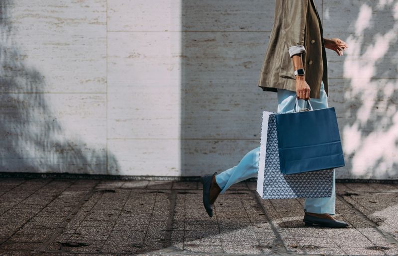 A stylish woman walking casually while carrying large shopping bags on a city sidewalk.