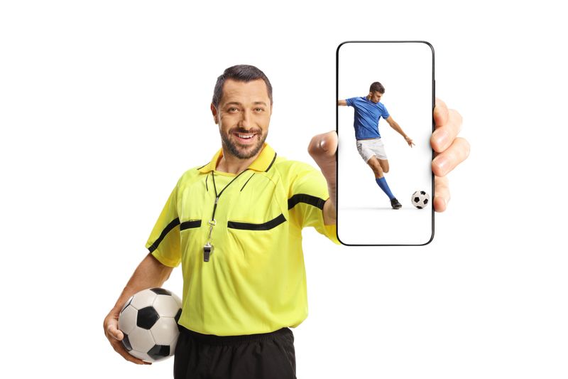 Football referee holding a ball and showing a smartphone with a football player on screen isolated on white background