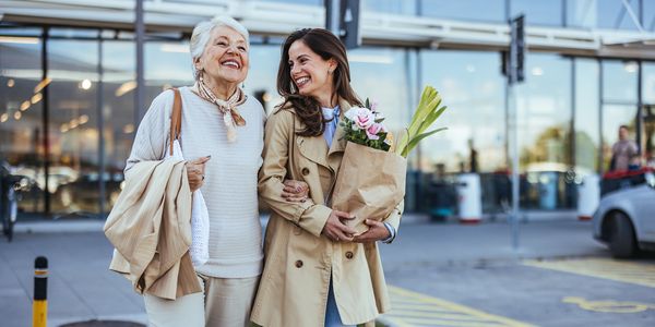Two women happily walking outside a store, carrying groceries and flowers.