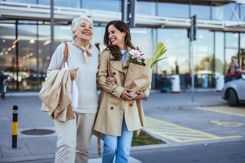 A joyful adult daughter walks with her elderly mother outside a store, both carrying grocery bags and sharing a light-hearted moment together.