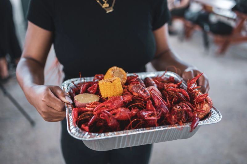 Close-up of a person holding a tray filled with cajun crawfish, corn, and potatoes at an outdoor gathering. The mix of seafood and vegetables suggests a casual and festive atmosphere, ideal for social events and culinary experiences.