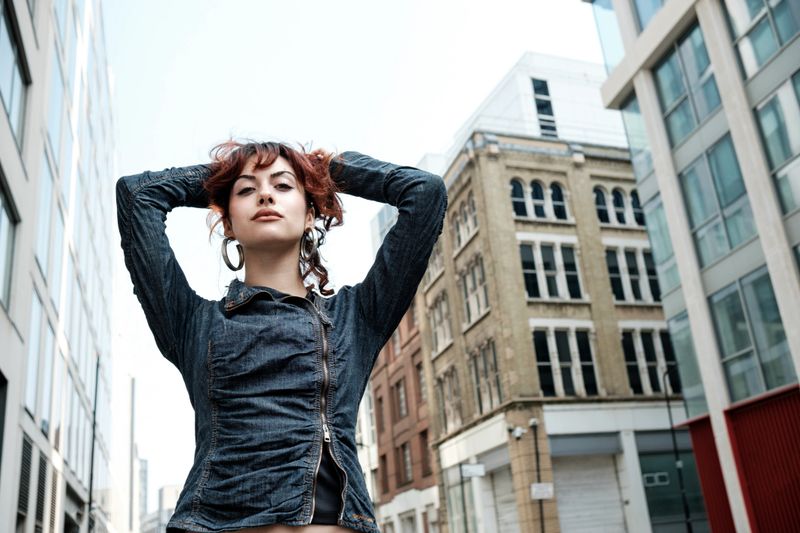 Low point of view of proud young woman with arms up on her head in the a London street. She is wearing a casual outfit, denim jacket and huge earrings. Urban vibes.