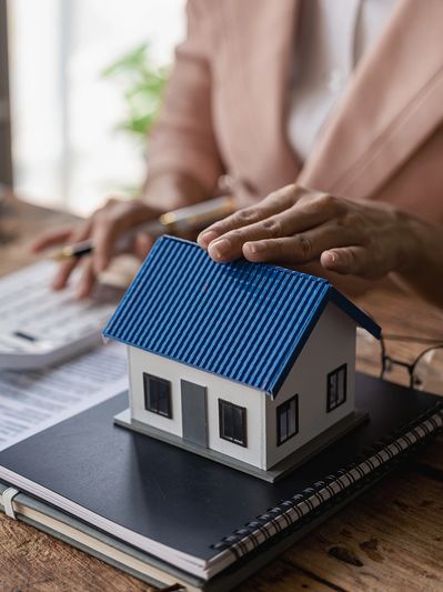 A woman using a calculator with a small model house, a laptop, and contract documents on a desk.