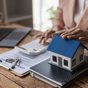 Person handling house model and paperwork with keys on table.