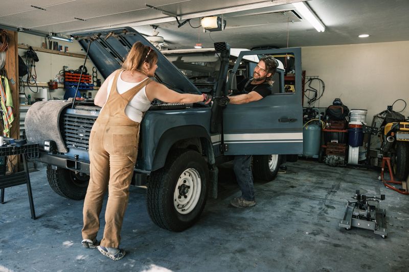 Mid adult UK immigrant auto mechanic couple working in their garage on a vintage 4x4 vehicle. Both dressed in work clothes. Interior of private garage of private home in Spring, Texas.