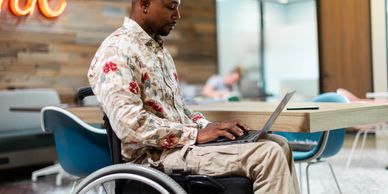 A black male wheelchair user in an office with a laptop on his lap.