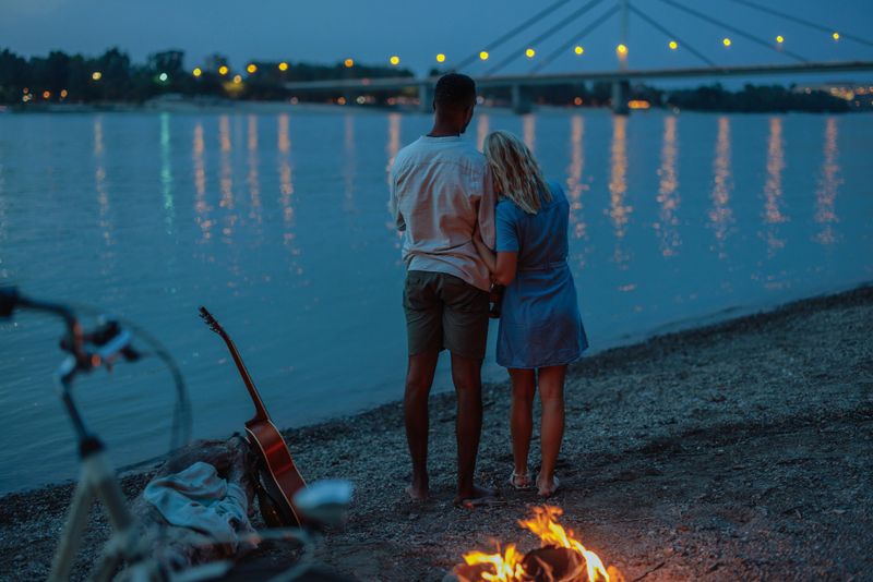 African-American guy and a Caucasian girl are hugging while standing on the river beach during the nightfall by the fire