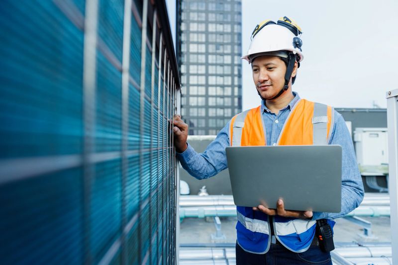 Man engineer holding laptop working at rooftop building construction. Male technician worker working checking hvac of office building. Engineering installing large air conditioning system.