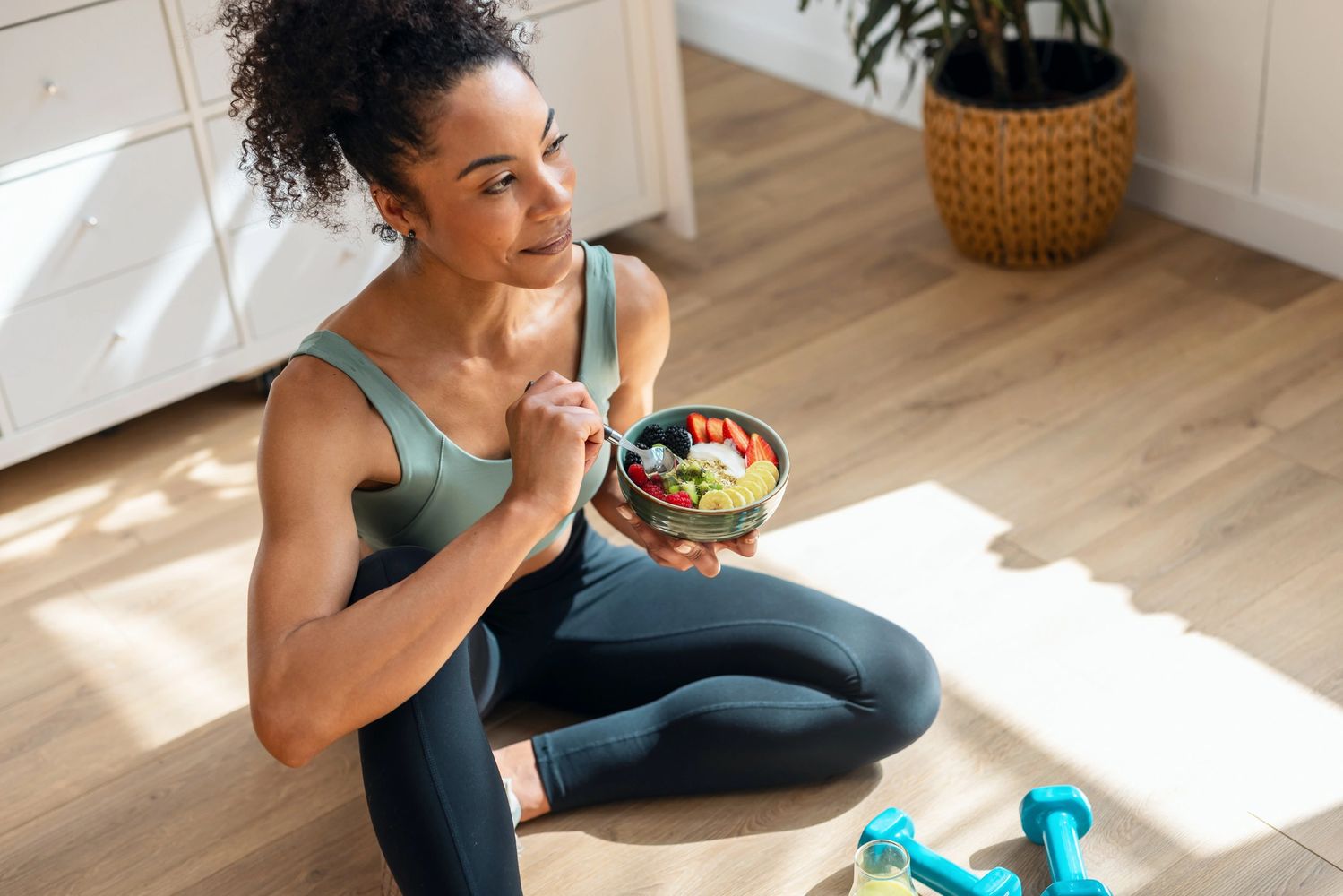 Woman in workout clothes enjoying a healthy fruit bowl while sitting on the floor.