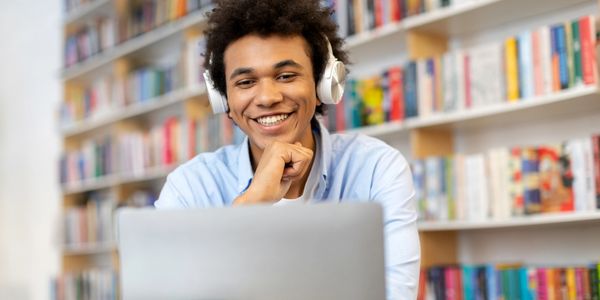 Young man with headphones smiling at a laptop in a library.