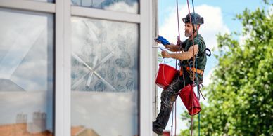 A man cleaning windows while suspended by ropes, wearing safety gear.