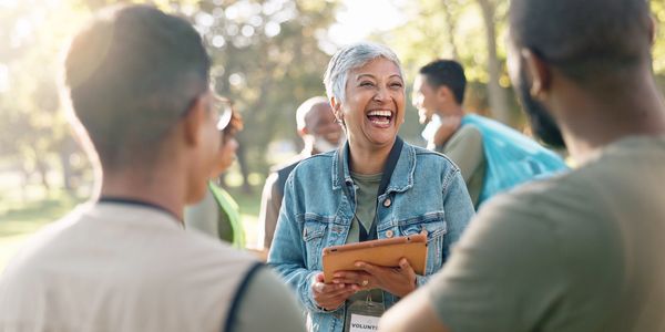 A joyful volunteer woman laughs while holding a tablet outdoors with a group of people.