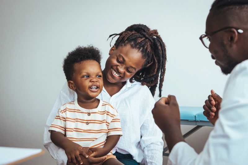 A joyful black boy sits on his mother's lap during a medical consultation as the doctor engages them in conversation, ensuring a soothing healthcare environment.