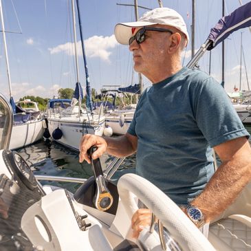 Older man steering a boat in a marina on a sunny day.