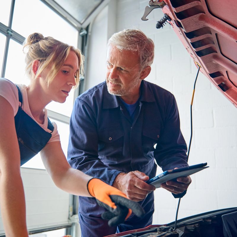 Male Car Auto Mechanic With Female Trainee Looking Under Bonnet Of Car Holding Digital Tablet