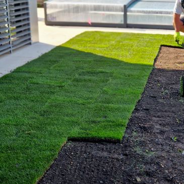 Workers laying fresh sod on a garden bed beside a building.