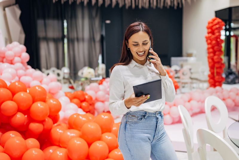 A decorator is standing in a restaurant surrounded by balloon decorations and talking on the phone with a new client to agree on a detailed plan for the decoration and organization of the party