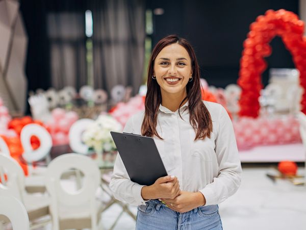 Smiling event planner holding a clipboard in a decorated venue.