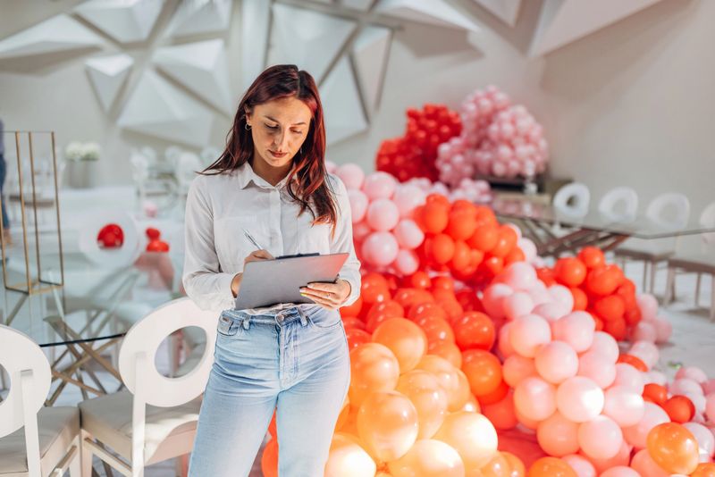 Female party organizer standing in restaurant surrounded by balloon decoration and holding clipboard