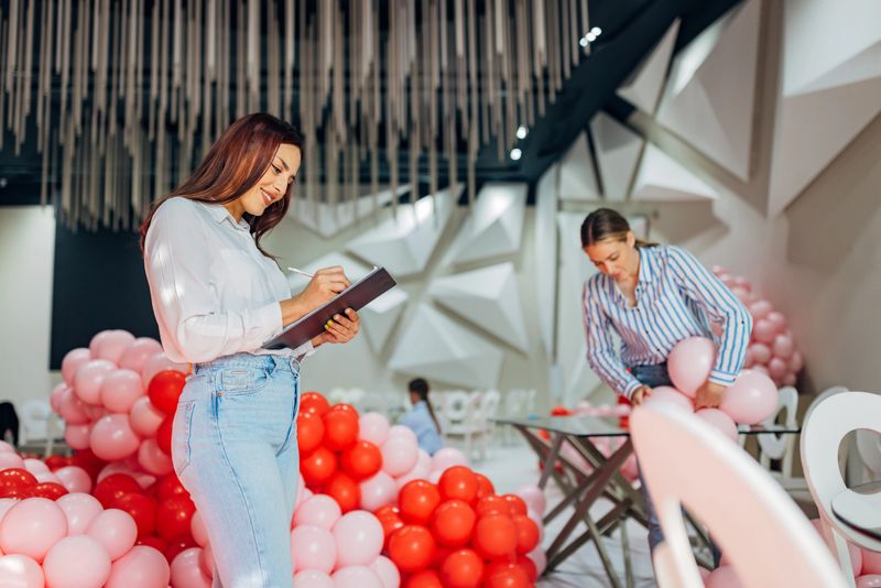 A woman party planner crosses tasks off her to-do list while decorating a restaurant for a party, while a decorator makes balloon decorations