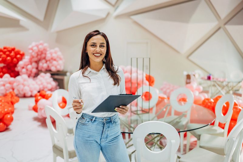 Waist up view of cheerful and beautiful female party organizer standing in restaurant surrounded by balloon decoration and holding clipboard