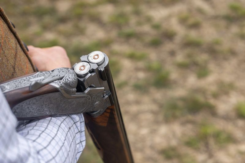 Close-up shot of an open shotgun barrel with shotgun cartridges in it. The man's arm is visible as the gun is hanging over it.