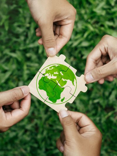 Three hands holding puzzle pieces forming a green Earth on a natural background.