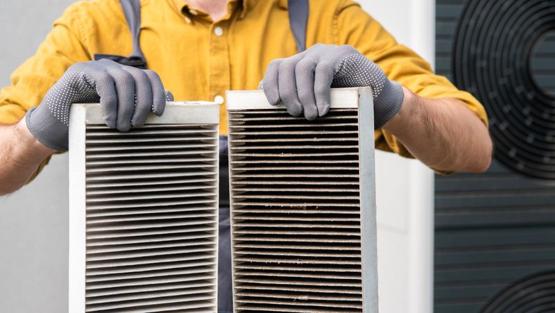 A worker in a yellow shirt and gloves stands outside, holding two dirty air filters from a home ventilation system. The filters show significant dirt, indicating the need for regular maintenance. The background includes part of the ventilation unit, emphasizing the importance of keeping the system clean.