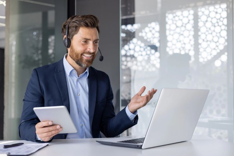 A professional businessman is engaging in a virtual meeting using a headset and laptop while holding a tablet in a modern office environment.