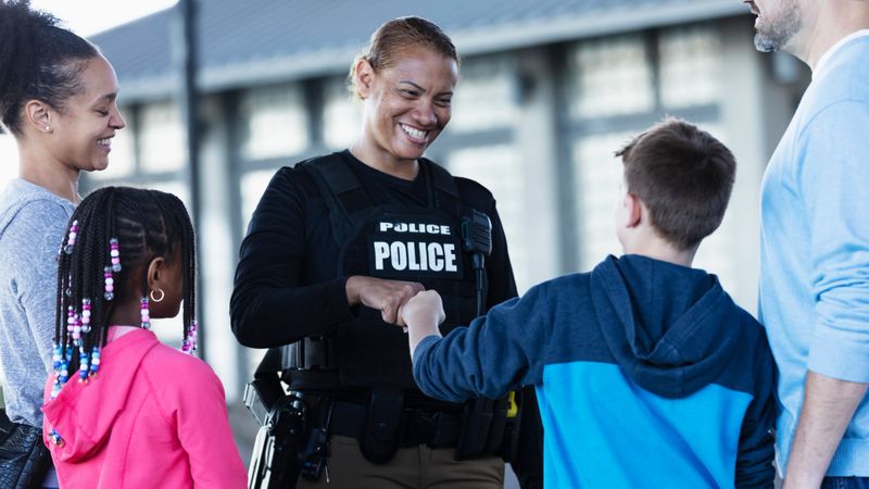 A multiracial police woman talking to a group of parents and children outdoors. standing outside a building. She is smiling at an 11 year old boy and they are bumping fists. The view is from over the boy's shoulder.