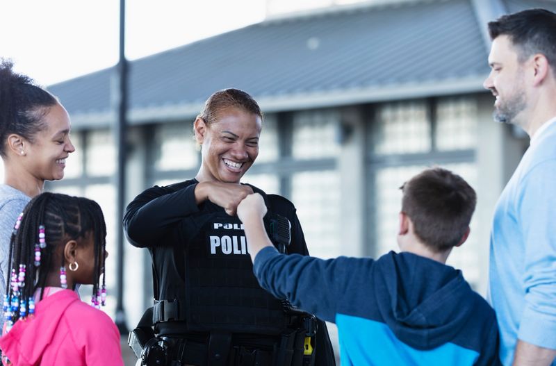 A multiracial police woman talking to a group of parents and children outdoors. standing outside a building. She is smiling at an 11 year old boy and they are bumping fists. The view is from over the boy's shoulder.