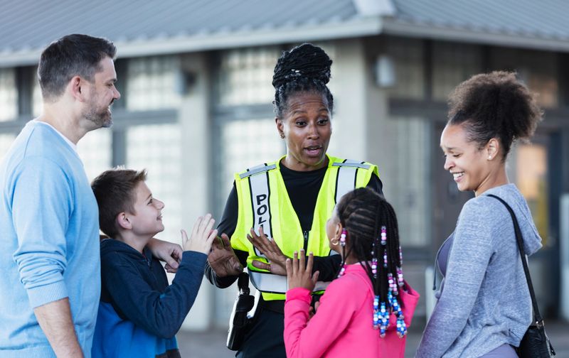 An African-American policewoman conversing with a multiracial group of parents and children outdoors. Everyone is smiling.