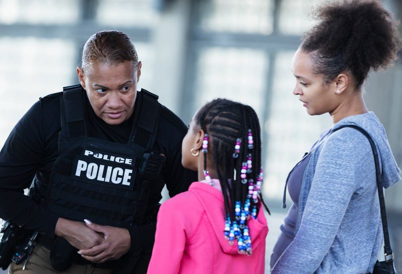 A multiracial policewomen, in her 40s, conversing with a 10 year old African-American girl and her mother. They are standing outdoors by a building. The view is from over the shoulder of the girl and the focus is on the police officer.