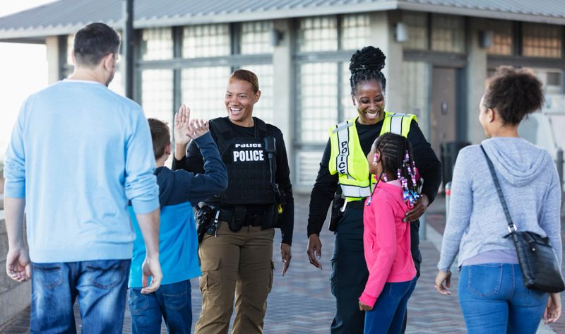 Two multiracial policewomen, in their 40s and 50s, conversing outdoors with two children and their parents. The view is from over the shoulders of the families, and the focus is on the police officers who are smiling at the children. One is giving a high five to the 11 year old boy.