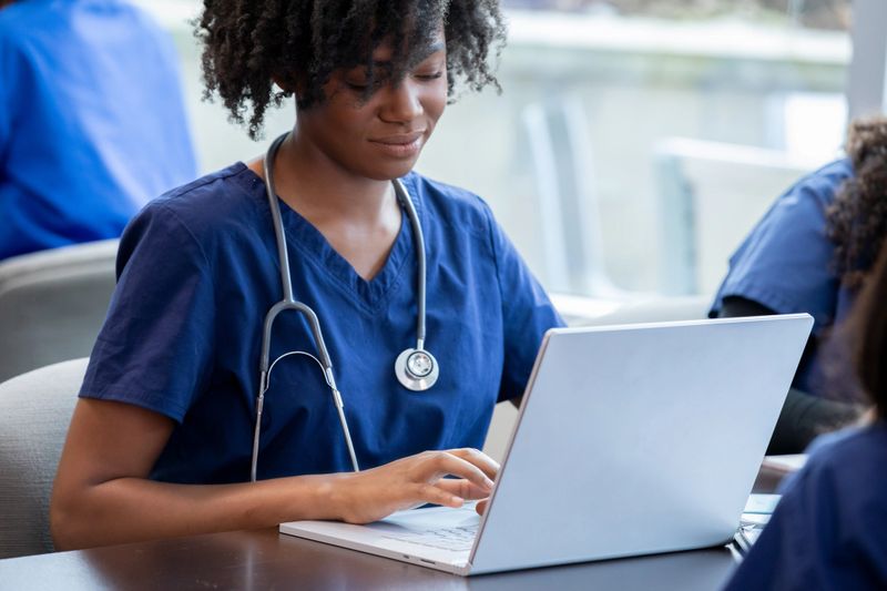 In class, a diligent female nursing student in scrubs engages in the discussion and takes notes on her laptop simultaneously.