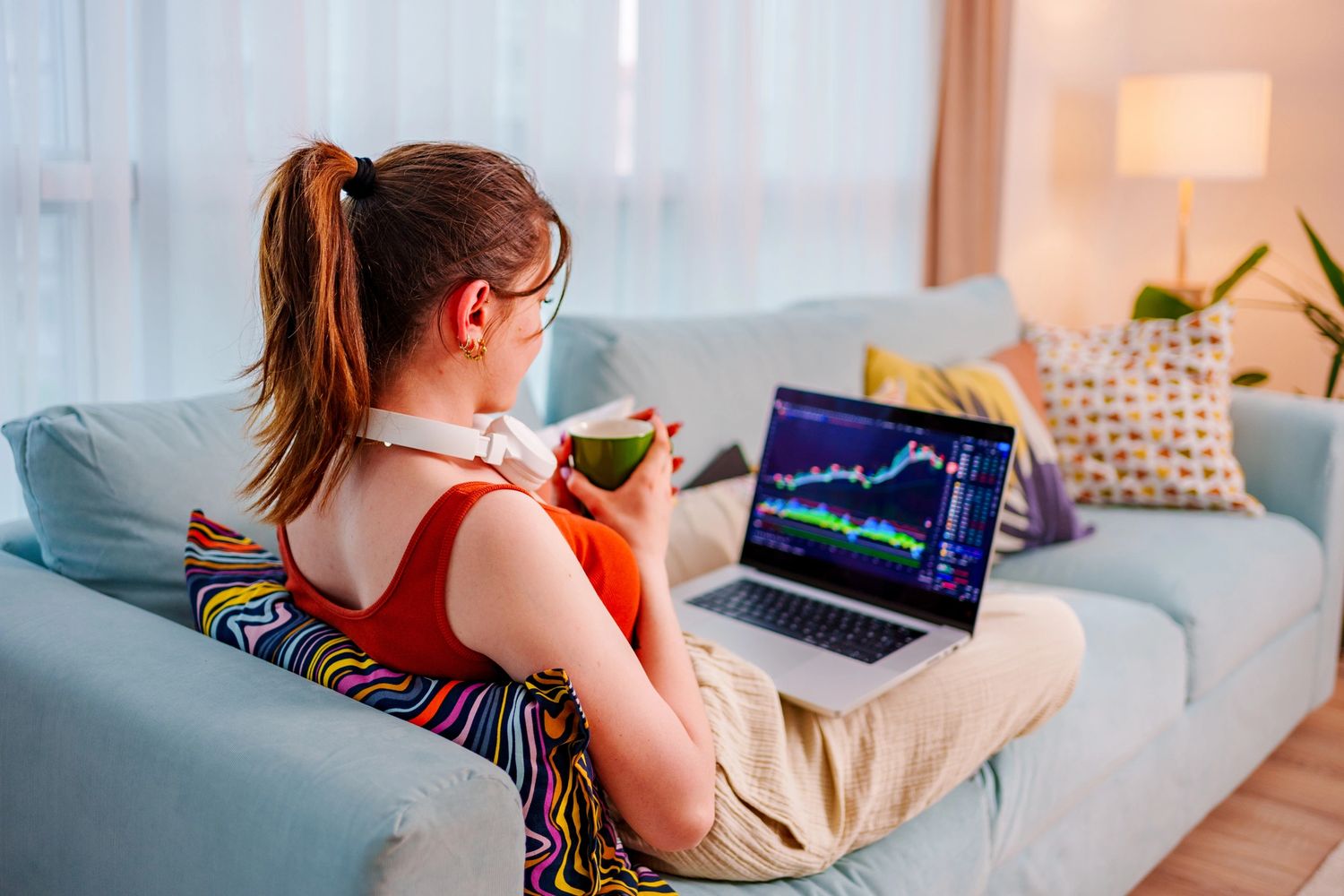 Woman relaxing on couch with coffee, analyzing stock charts on laptop.