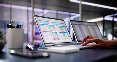 Two laptops displaying spreadsheets on a desk with a hand using one laptop.