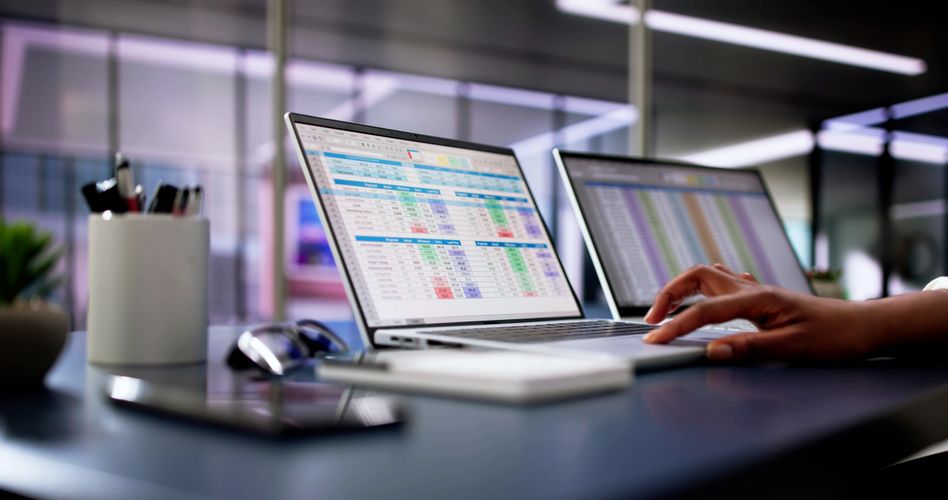 Person working on financial spreadsheets on two laptops in an office.