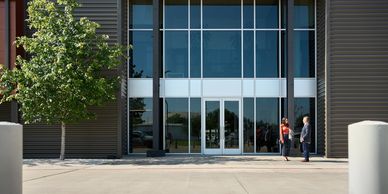 Two people stand outside a modern building entrance with large glass doors.