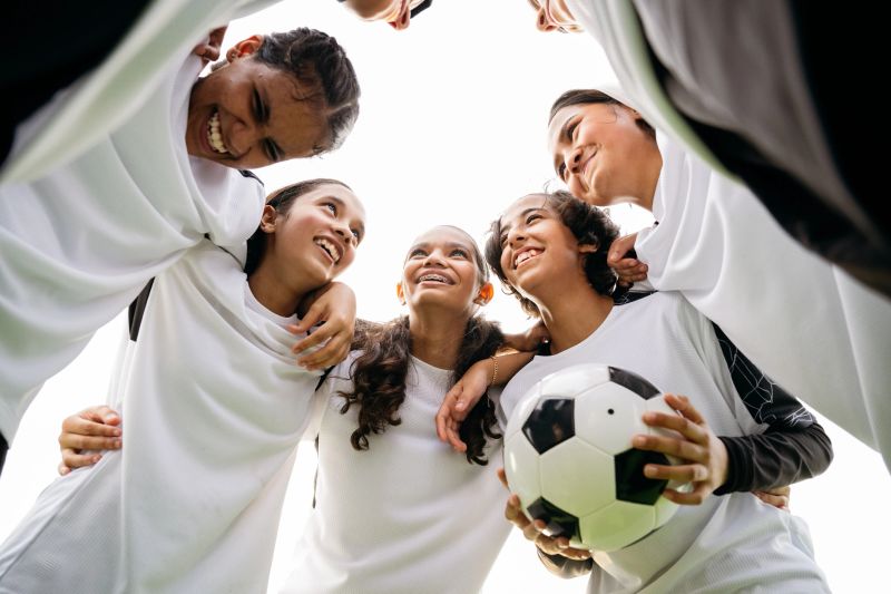 Low angle view of cheerful soccer players standing in huddle with ball. Female athletes are with arms around each other. They are planning strategy together.