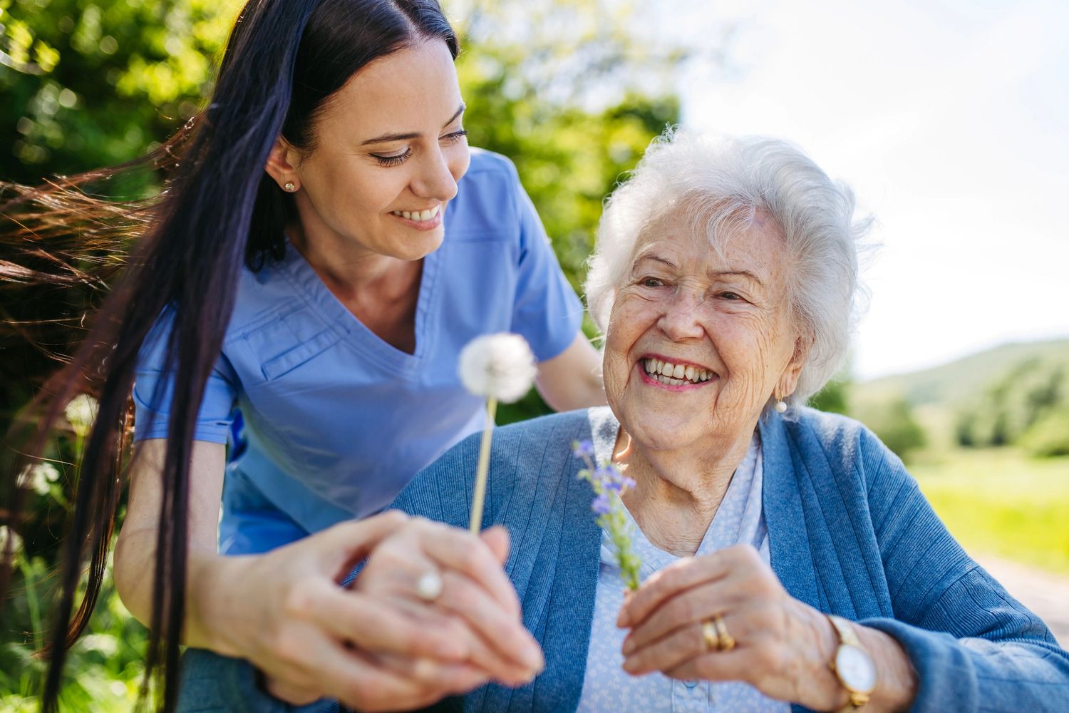 A young caregiver and elderly woman share a joyful moment outdoors with flowers.