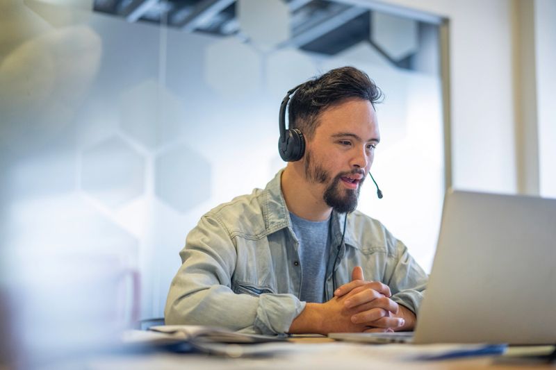 Medium front view shot of young man wearing headset while participating in video conference call at inclusive office