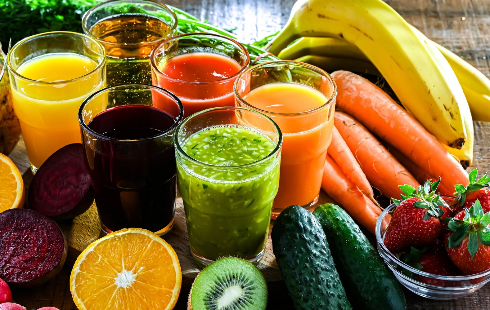 A variety of fresh fruit and vegetable juices surrounded by colorful produce on a wooden table.