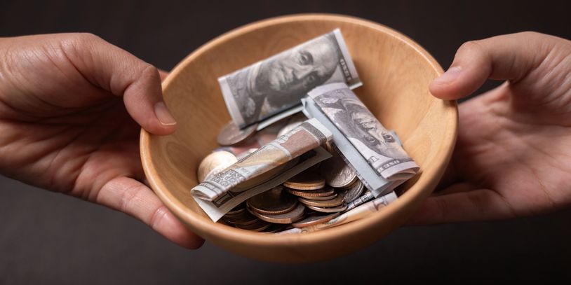 Hands holding a wooden bowl filled with coins and folded dollar bills.
