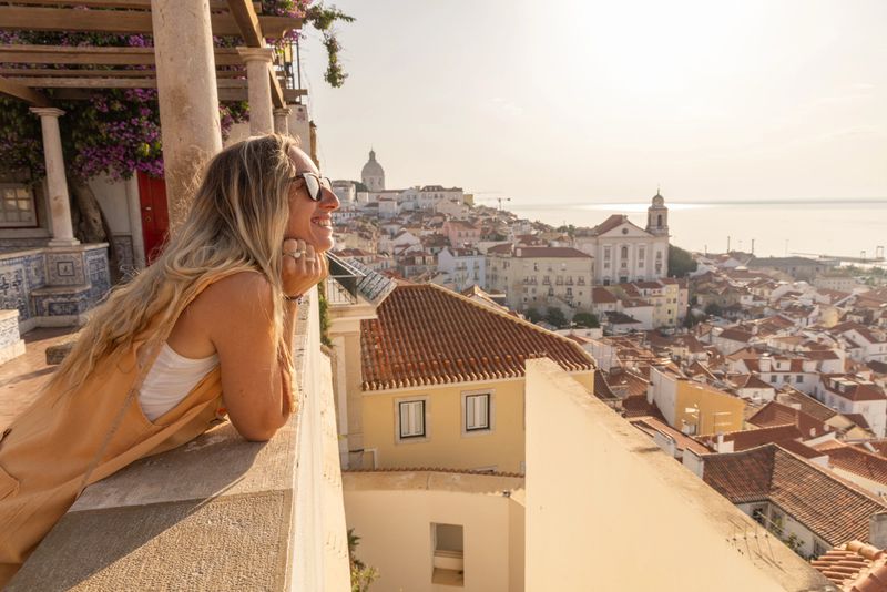 A smiling woman with blonde hair and sunglasses leans on a traditional terrace, gazing at the scenic rooftops and ocean view of Lisbon's Alfama district in the warm morning light.