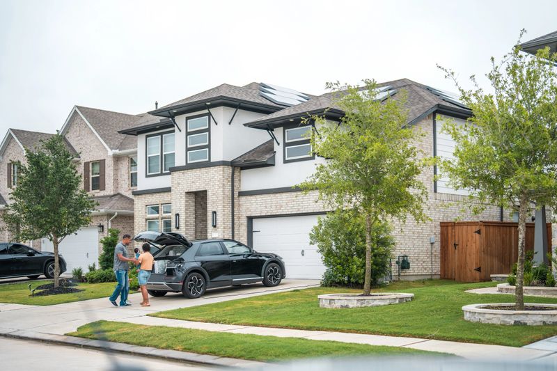 A multiracial family engages in unloading shopping bags near an electric car, with a solar-paneled house in the serene background.